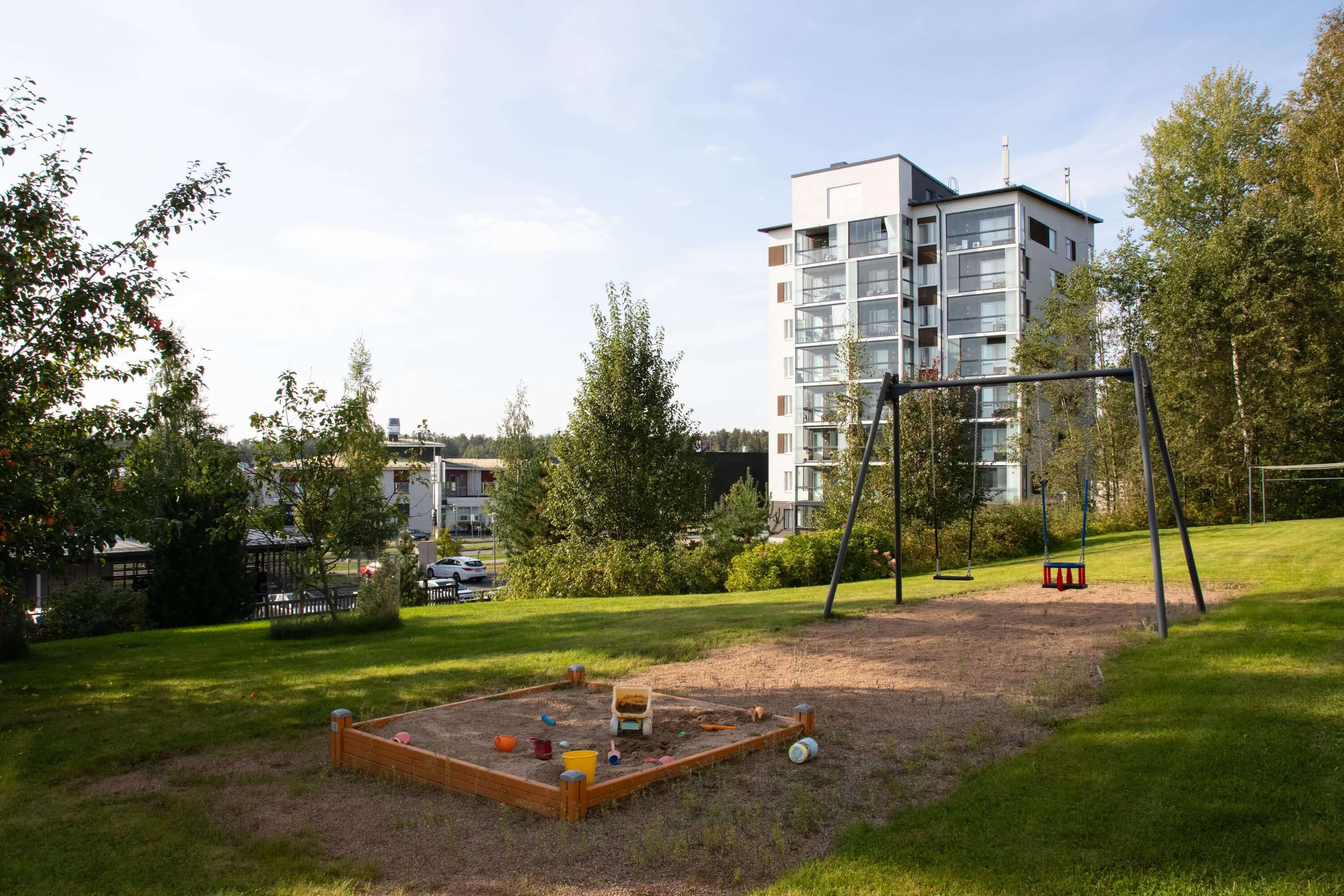 Asuntokuvaus: leikkipaikka ja asuinrakennus vehreässä ympäristössä, lasten leikkivälineet ja puut.