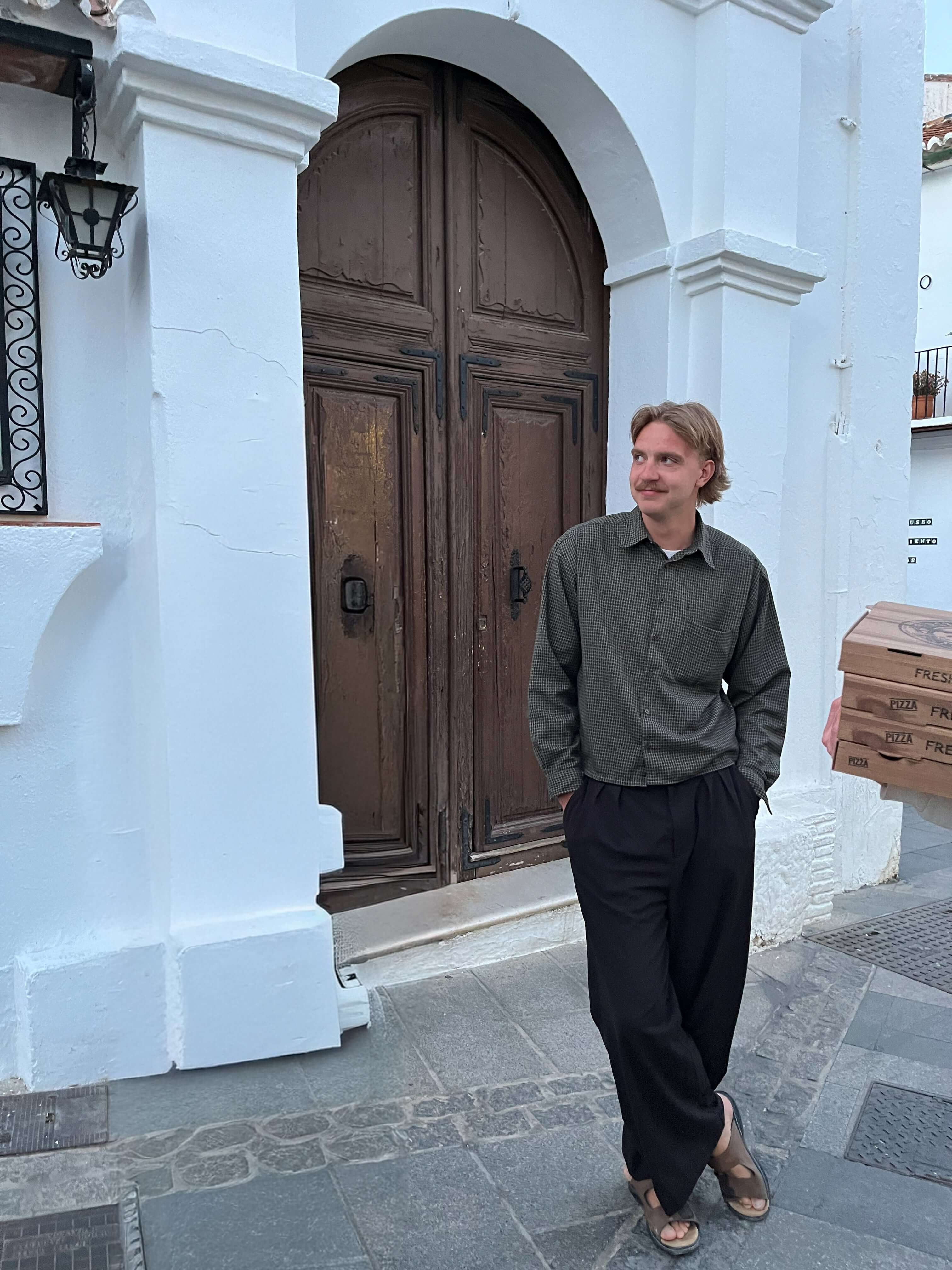 A person casually standing in front of a classic wooden door, wearing a dark shirt and black pants.